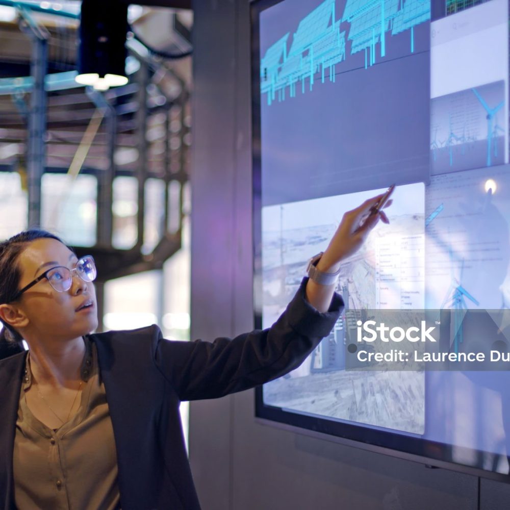 Stock photograph of a young Asian woman conducting a seminar / lecture with the aid of a large screen. The screen is displaying data & designs concerning low carbon electricity production with solar panels & wind turbines. These are juxtaposed with an image of conventional fossil fuel oil production.